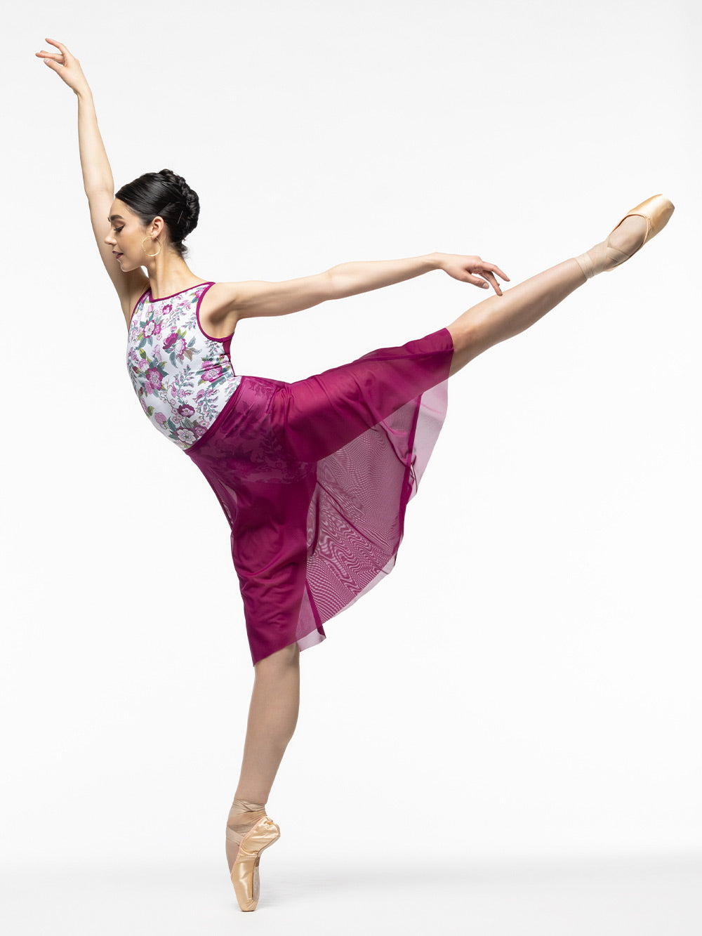Model is wearing white, magenta, and green floral print leotard with open strappy back and magenta trim and mesh panels. Paired with long magenta mesh skirt.