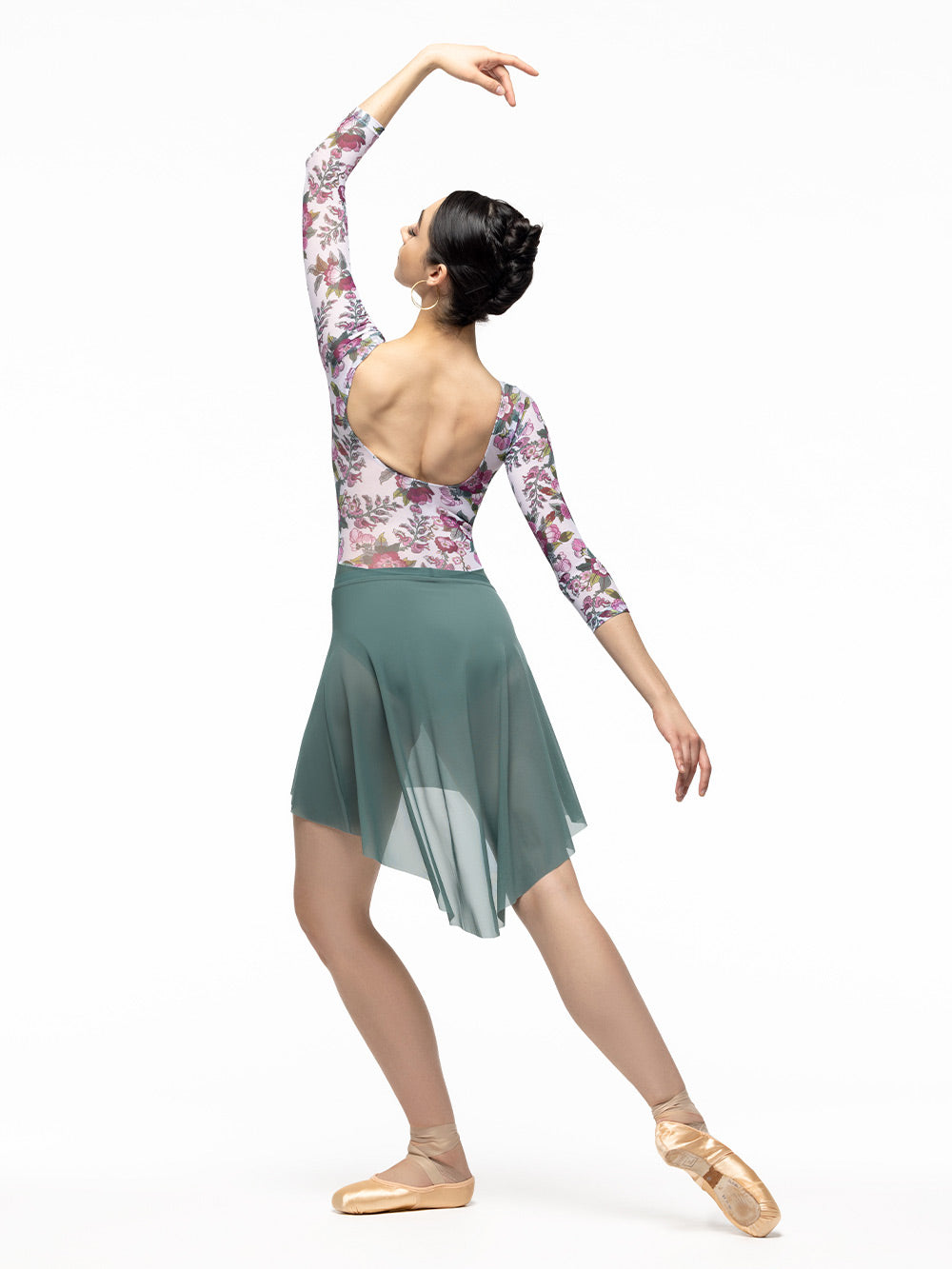 Back of model wearing pale green leotard with white, magenta, and green floral print mesh top, scoop back, and below elbow sleeves. Paired with pale green high low mesh skirt.