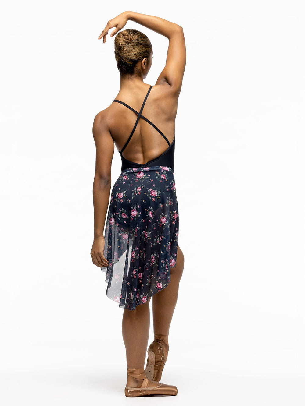Back of model wearing solid black leotard with front and back pinch and crossed cami straps. Paired with dark navy and pink floral print high low mesh skirt.