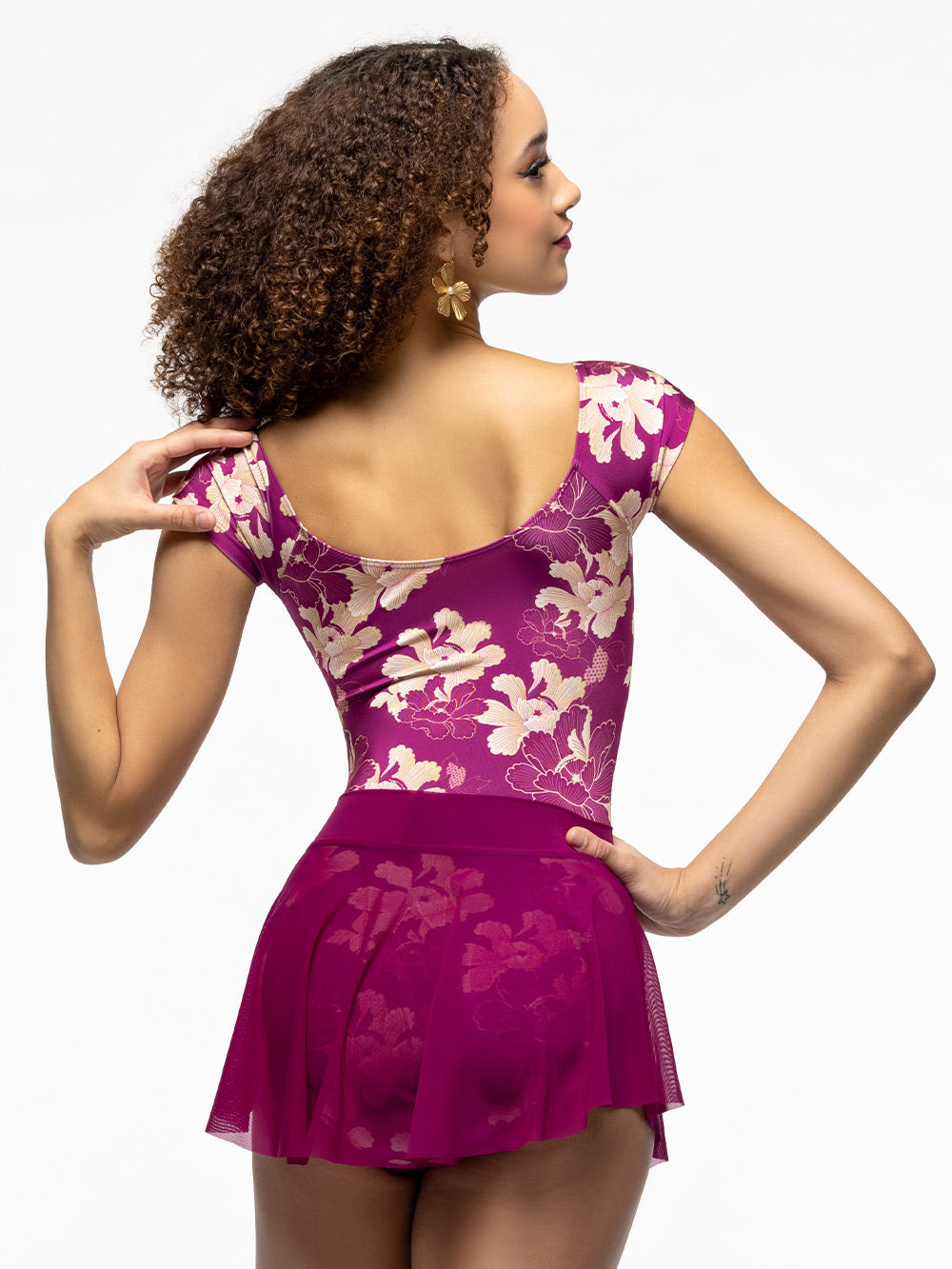 Back of model wearing shiny magenta and gold floral print leotard with cap sleeves, square neck, and scoop back. Paired with dark magenta mesh hipster skirt.