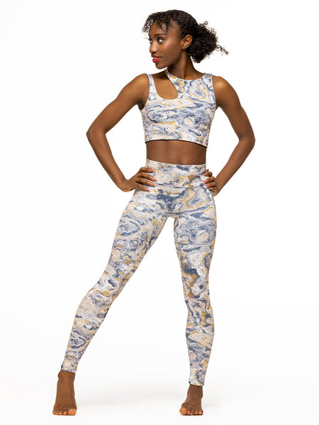 Model is wearing white, gold, and navy blue ink swirl print high waisted leggings with matching activewear crop top with a shoulder cutout.