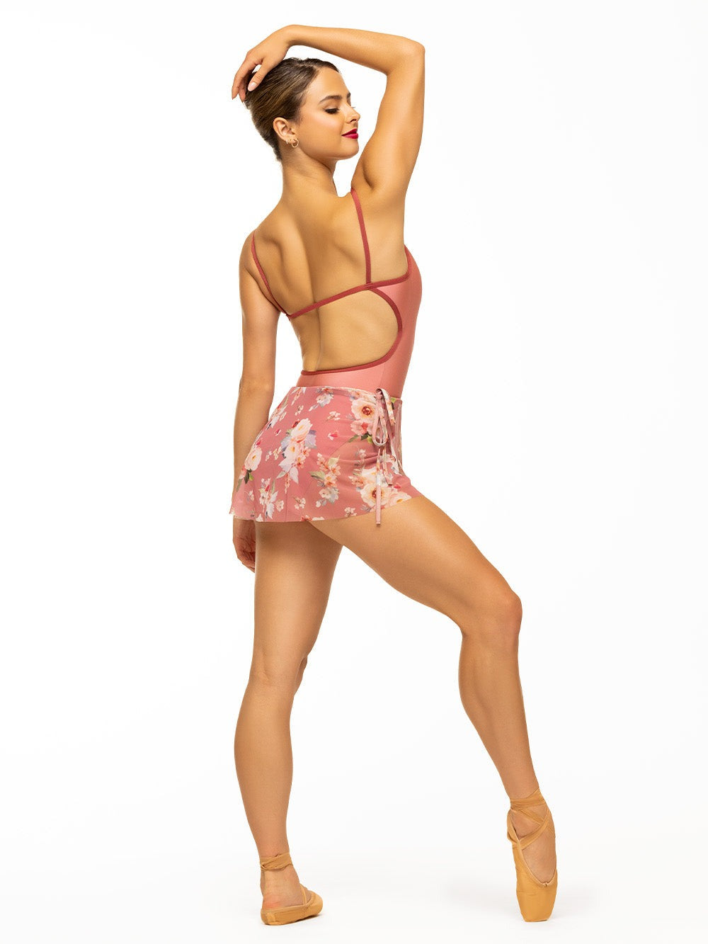 Back of model wearing shiny pink bodysuit with rust trim and sporty open back paired with pink floral print mesh mini wrap skirt.