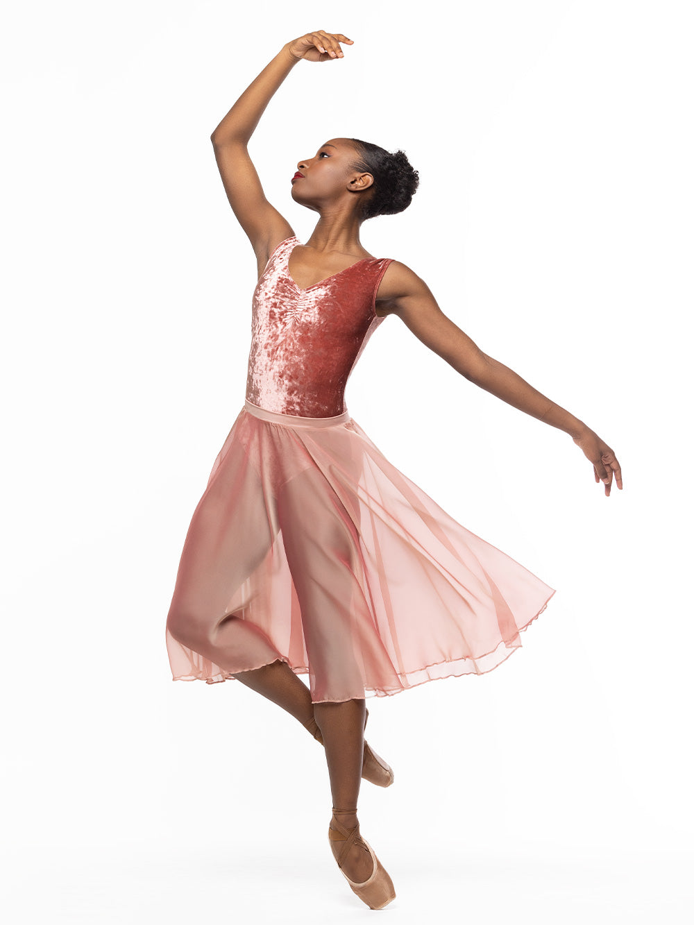 Dancer is on point with cou-de-pie derriere facing diagonal front with one arm on fifth position, wearing a Chiffon Rehearsal Skirt Sandstone RTW.