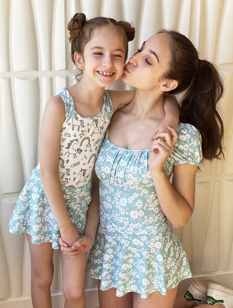 two models, one wearing a unicorn pattern leotard with blue floral straps and skirt, Model two wearing a blue floral leotard with matching skirt