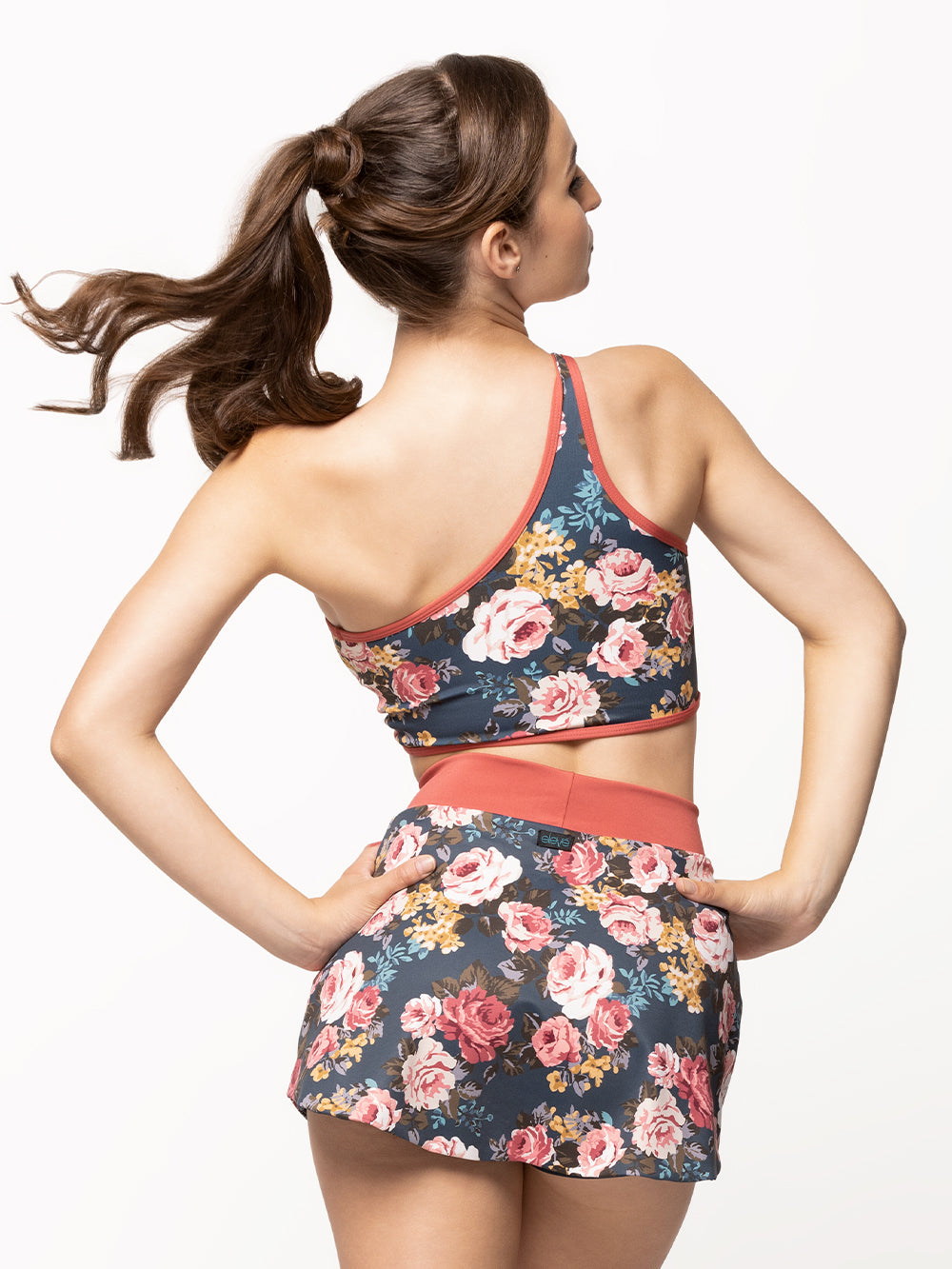 Back of model wearing a navy blue floral print tennis skirt with a mineral red waistband and attached shorts with a matching one-shoulder crop top