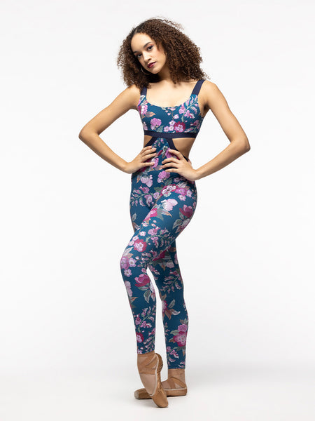Model is wearing teal, magenta, and green floral print full length unitard with navy blue elastic straps and a side to back cutout.